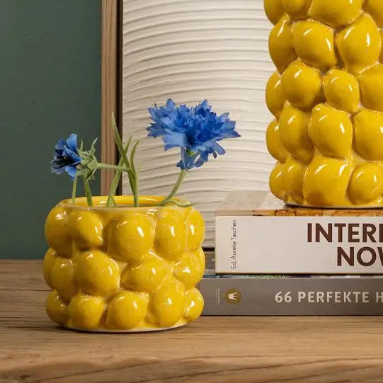 Yellow ceramic vase with floral arrangement on a wooden surface with a framed picture in the background.